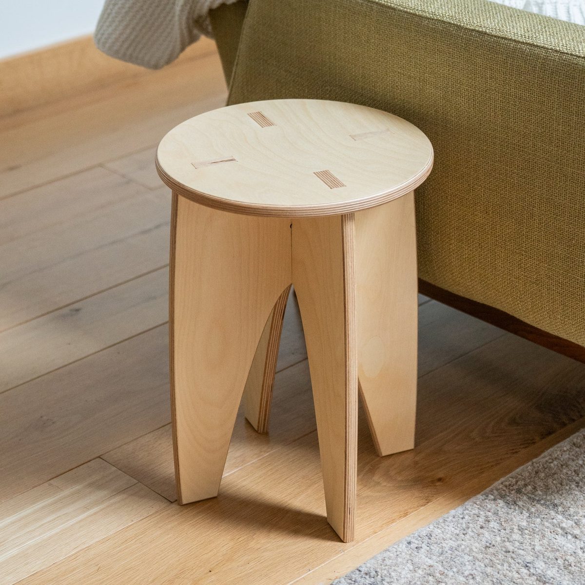 The assembled plywood stool on a wood floor beside a sofa, with four tab ends showing flush in the round seat.