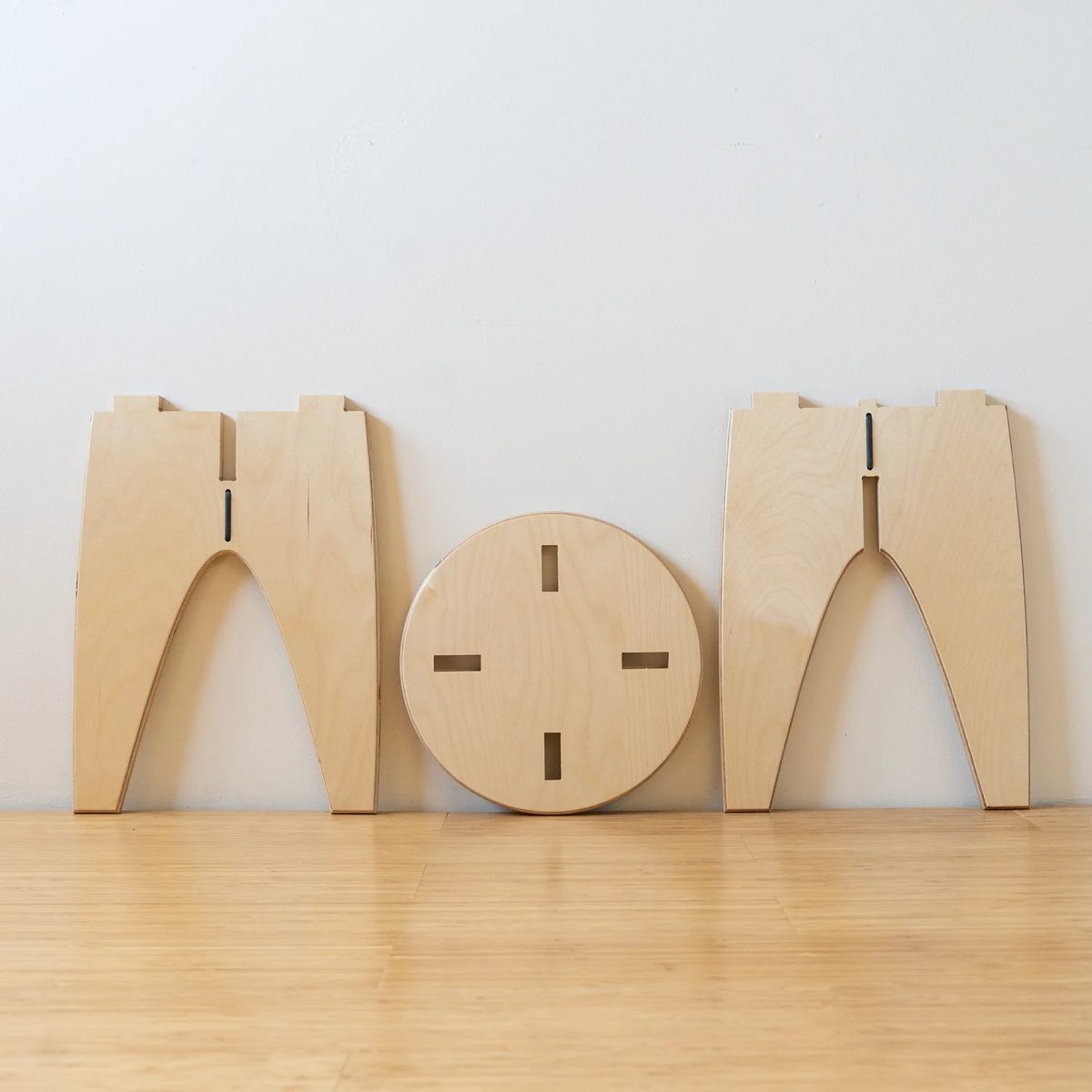 Two plywood leg panels with long central slots and a round plywood seat with four rectangular holes, leaning against a wall.